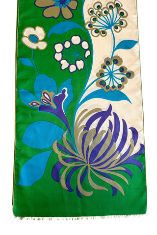 A vintage 1960s scarf by designer Jean Dessès made of silk twill, with a large colourblock grass green and cream background, with a large stylized floral print featuring approximately six large different types of flowers in combinations of turquoise blue, indigo, cream, and beige colours. The name of the designer Jean Dessès is printed at the bottom right of the long rectangular scarf.