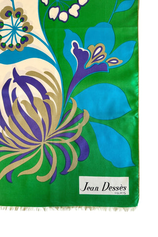 A vintage 1960s scarf by designer Jean Dessès made of silk twill, with a large colourblock grass green and cream background, with a large stylized floral print featuring approximately six large different types of flowers in combinations of turquoise blue, indigo, cream, and beige colours. The name of the designer Jean Dessès is printed at the bottom right of the long rectangular scarf.