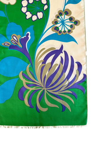 A vintage 1960s scarf by designer Jean Dessès made of silk twill, with a large colourblock grass green and cream background, with a large stylized floral print featuring approximately six large different types of flowers in combinations of turquoise blue, indigo, cream, and beige colours. The name of the designer Jean Dessès is printed at the bottom right of the long rectangular scarf.