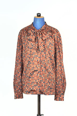 Silky smooth light brown blouse with blue, orange, and rust polkadot print. Features a pussy bow collar, slightly gathered shoulders and covered buttons. The sleeves have some volume at the buttoned cuffs. 