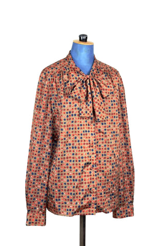 Silky smooth light brown blouse with blue, orange, and rust polkadot print. Features a pussy bow collar, slightly gathered shoulders and covered buttons. The sleeves have some volume at the buttoned cuffs. 