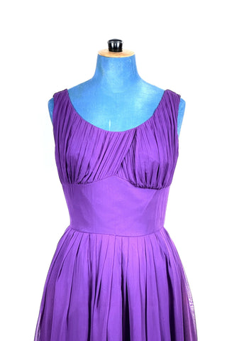 Show-stopping vintage 1950s party dress in bright purple chiffon. The voluminous skirt is pleated at the waist, echoing the gathered draped bust. The wide scoop neckline highlights your décolletage. Lined in purple chiffon, with a reinforced cotton inset waist lining for extra structure. Closes in the back with a metal zipper.