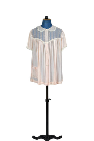 Charming vintage 1950s bed jacket pajama top in silky pink acetate & rayon by Barbizon “Lazy Lizzie”. Beautifully constructed with an inset yoke panel of sheer acetate with delicate pintucks, a Peter Pan collar edged with gathered lace trim, and short sleeves also trimmed in lace. Closes with four mother-of-pearl buttons. 