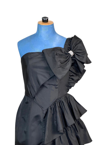 Incredible vintage 1980s cocktail dress in black rayon with tiered ruffles at the shoulder and down the left hip. A giant bow is draped at the shoulder, finished with a sparkling band of rhinestones. The 1940s-inspired draping and fit of this dress are impeccable, with a nipped waist and figure-hugging bodice and skirt. Closes with a zipper and hook-and-eye closure at the back. 