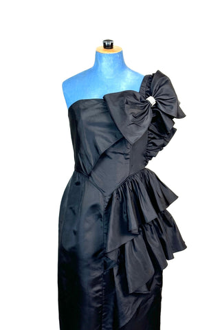 Incredible vintage 1980s cocktail dress in black rayon with tiered ruffles at the shoulder and down the left hip. A giant bow is draped at the shoulder, finished with a sparkling band of rhinestones. The 1940s-inspired draping and fit of this dress are impeccable, with a nipped waist and figure-hugging bodice and skirt. Closes with a zipper and hook-and-eye closure at the back. 