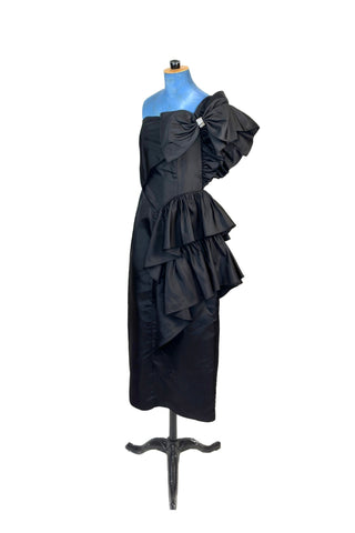 Incredible vintage 1980s cocktail dress in black rayon with tiered ruffles at the shoulder and down the left hip. A giant bow is draped at the shoulder, finished with a sparkling band of rhinestones. The 1940s-inspired draping and fit of this dress are impeccable, with a nipped waist and figure-hugging bodice and skirt. Closes with a zipper and hook-and-eye closure at the back. 