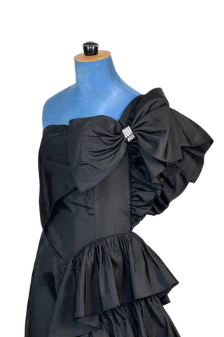 Incredible vintage 1980s cocktail dress in black rayon with tiered ruffles at the shoulder and down the left hip. A giant bow is draped at the shoulder, finished with a sparkling band of rhinestones. The 1940s-inspired draping and fit of this dress are impeccable, with a nipped waist and figure-hugging bodice and skirt. Closes with a zipper and hook-and-eye closure at the back. 