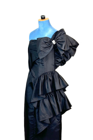 Incredible vintage 1980s cocktail dress in black rayon with tiered ruffles at the shoulder and down the left hip. A giant bow is draped at the shoulder, finished with a sparkling band of rhinestones. The 1940s-inspired draping and fit of this dress are impeccable, with a nipped waist and figure-hugging bodice and skirt. Closes with a zipper and hook-and-eye closure at the back. 