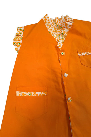Bright tangerine orange reversible vintage 1960s girls’ vest with orange and green floral print. Each reversible side has pockets trimmed in the reverse-side fabric. The neckline and sleeve holes are trimmed with self-fabric ruffles.