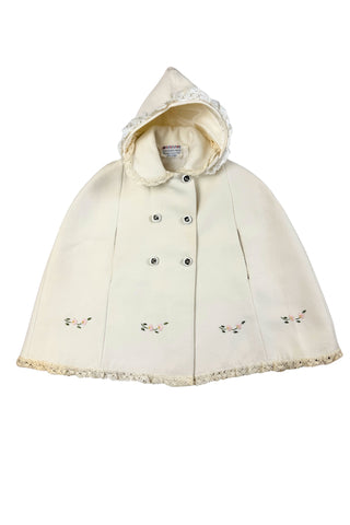 This darling vintage 1960s cape and dress set is embroidered with pink cherry blossoms and pink, blue and yellow tulips. The cape has a detachable hood which attaches with buttons at the back and sides of the cape’s round Peter Pan collar. The collar and hem of the cape are trimmed with scalloped crocheted lace. The cape has a double-breasted front closure with white and silver tone square buttons. There are arm holes in the middle of the front seams.