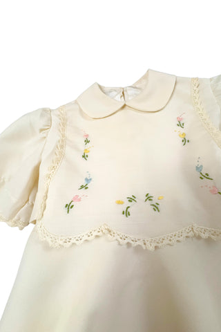 This darling vintage 1960s cape and dress set is embroidered with pink cherry blossoms and pink, blue and yellow tulips. The cape has a detachable hood which attaches with buttons at the back and sides of the cape’s round Peter Pan collar. The collar and hem of the cape are trimmed with scalloped crocheted lace. The cape has a double-breasted front closure with white and silver tone square buttons. There are arm holes in the middle of the front seams.