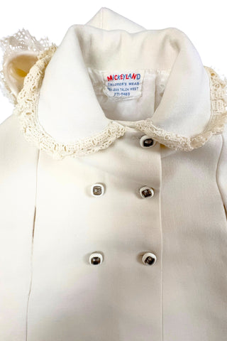 This darling vintage 1960s cape and dress set is embroidered with pink cherry blossoms and pink, blue and yellow tulips. The cape has a detachable hood which attaches with buttons at the back and sides of the cape’s round Peter Pan collar. The collar and hem of the cape are trimmed with scalloped crocheted lace. The cape has a double-breasted front closure with white and silver tone square buttons. There are arm holes in the middle of the front seams.