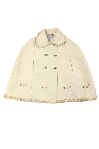 This darling vintage 1960s cape and dress set is embroidered with pink cherry blossoms and pink, blue and yellow tulips. The cape has a detachable hood which attaches with buttons at the back and sides of the cape’s round Peter Pan collar. The collar and hem of the cape are trimmed with scalloped crocheted lace. The cape has a double-breasted front closure with white and silver tone square buttons. There are arm holes in the middle of the front seams.