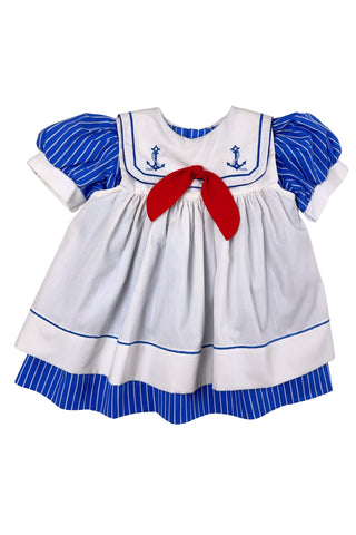 Adorable vintage 1980s toddler sailor dress with separate blue and white striped under-dress and white bib-collar pinafore apron which closes in the back with blue and white buttons. The sailor collar of the dress is embroidered with two anchors and finished with a bright red bow tie, while the pinafore has a matching blue stripe around the hem. The dress closes in the back with two buttons.
