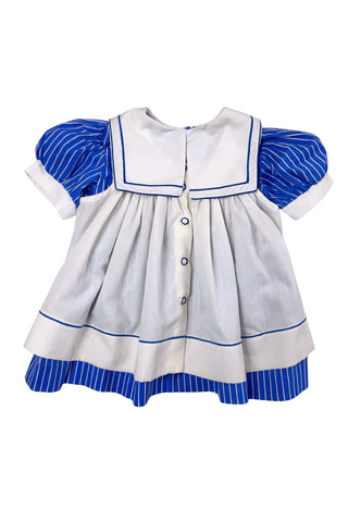 Adorable vintage 1980s toddler sailor dress with separate blue and white striped under-dress and white bib-collar pinafore apron which closes in the back with blue and white buttons. The sailor collar of the dress is embroidered with two anchors and finished with a bright red bow tie, while the pinafore has a matching blue stripe around the hem. The dress closes in the back with two buttons.