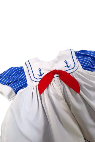 Adorable vintage 1980s toddler sailor dress with separate blue and white striped under-dress and white bib-collar pinafore apron which closes in the back with blue and white buttons. The sailor collar of the dress is embroidered with two anchors and finished with a bright red bow tie, while the pinafore has a matching blue stripe around the hem. The dress closes in the back with two buttons.