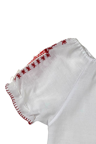 Vintage 1970s peasant blouse in white cotton with red hand embroidery stitching and red and white floral appliqué embroidered patches on the chest and sleeves. The neckline and sleeves are gathered with tiny white cord drawstring. The edges of the sleeves are trimmed with red blanket stitch.