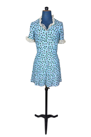 Vintage 1960s mini dress in sky blue with green and white floral print. The V neck and short sleeves are trimmed with wide white lace ruffles. The dress has an inset waistband, with slightly gathered bust. The A-line skirt hits at mid-thigh. The dress closes in the back with a cotton twill zipper and bow sash tie, and is unlined.