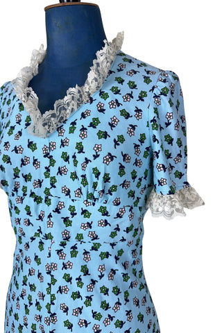 Vintage 1960s mini dress in sky blue with green and white floral print. The V neck and short sleeves are trimmed with wide white lace ruffles. The dress has an inset waistband, with slightly gathered bust. The A-line skirt hits at mid-thigh. The dress closes in the back with a cotton twill zipper and bow sash tie, and is unlined.
