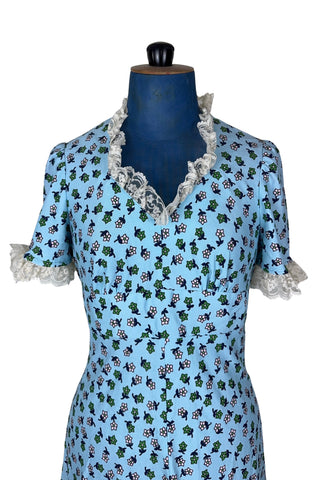 Vintage 1960s mini dress in sky blue with green and white floral print. The V neck and short sleeves are trimmed with wide white lace ruffles. The dress has an inset waistband, with slightly gathered bust. The A-line skirt hits at mid-thigh. The dress closes in the back with a cotton twill zipper and bow sash tie, and is unlined.