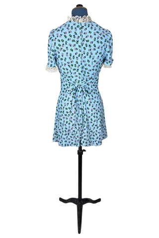 Vintage 1960s mini dress in sky blue with green and white floral print. The V neck and short sleeves are trimmed with wide white lace ruffles. The dress has an inset waistband, with slightly gathered bust. The A-line skirt hits at mid-thigh. The dress closes in the back with a cotton twill zipper and bow sash tie, and is unlined.