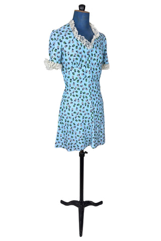Vintage 1960s mini dress in sky blue with green and white floral print. The V neck and short sleeves are trimmed with wide white lace ruffles. The dress has an inset waistband, with slightly gathered bust. The A-line skirt hits at mid-thigh. The dress closes in the back with a cotton twill zipper and bow sash tie, and is unlined.