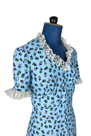 Vintage 1960s mini dress in sky blue with green and white floral print. The V neck and short sleeves are trimmed with wide white lace ruffles. The dress has an inset waistband, with slightly gathered bust. The A-line skirt hits at mid-thigh. The dress closes in the back with a cotton twill zipper and bow sash tie, and is unlined.