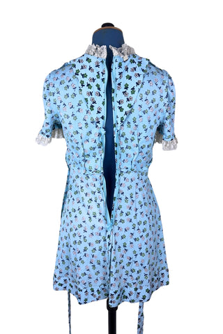 Vintage 1960s mini dress in sky blue with green and white floral print. The V neck and short sleeves are trimmed with wide white lace ruffles. The dress has an inset waistband, with slightly gathered bust. The A-line skirt hits at mid-thigh. The dress closes in the back with a cotton twill zipper and bow sash tie, and is unlined.