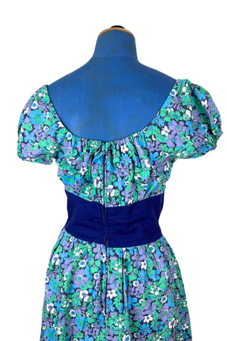 Vintage 1960s mini dress with laced bodice inspired by Alpine dirndl. The dress features a wide gathered neckline and puff sleeves, and closes in the back with a cotton twill zipper. It is made from green and blue floral print cotton with a wide navy blue bodice with silver crest buttons. The dress is unlined.