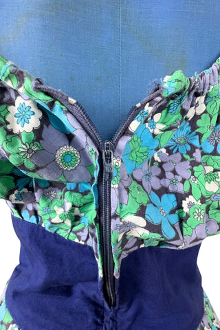 Vintage 1960s mini dress with laced bodice inspired by Alpine dirndl. The dress features a wide gathered neckline and puff sleeves, and closes in the back with a cotton twill zipper. It is made from green and blue floral print cotton with a wide navy blue bodice with silver crest buttons. The dress is unlined.