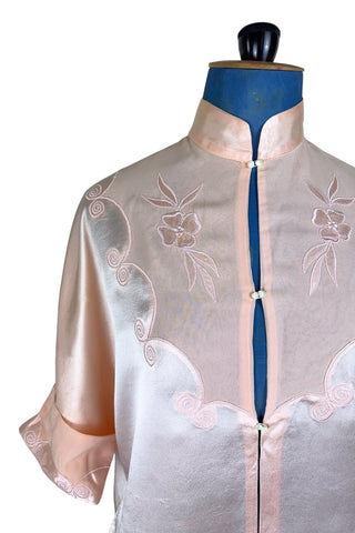 Vintage 1930s silk bed jacket with mandarin collar in mixed satin and crepe silk, with embroidered appliqué flowers and swirls. The three-quarter length sleeves have wide folded cuffs embellished with matching embroidered swirls. Closes in the front with three sets of tiny shiny buttons that resemble ivory.

Label reads Classic Silk Underwear. Made in Canada. Size Small.