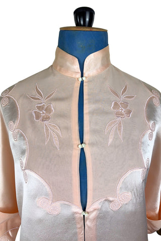 Vintage 1930s silk bed jacket with mandarin collar in mixed satin and crepe silk, with embroidered appliqué flowers and swirls. The three-quarter length sleeves have wide folded cuffs embellished with matching embroidered swirls. Closes in the front with three sets of tiny shiny buttons that resemble ivory.

Label reads Classic Silk Underwear. Made in Canada. Size Small.