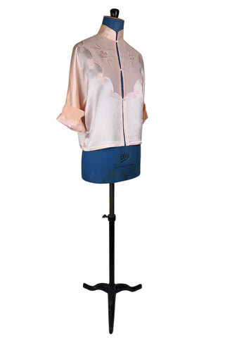Vintage 1930s silk bed jacket with mandarin collar in mixed satin and crepe silk, with embroidered appliqué flowers and swirls. The three-quarter length sleeves have wide folded cuffs embellished with matching embroidered swirls. Closes in the front with three sets of tiny shiny buttons that resemble ivory.

Label reads Classic Silk Underwear. Made in Canada. Size Small.