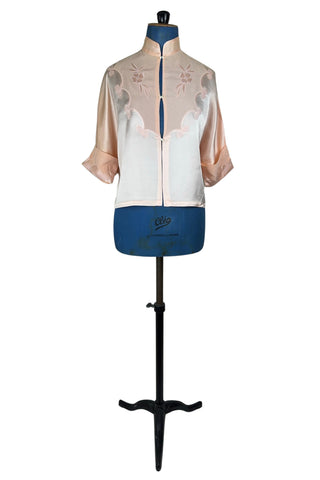 Vintage 1930s silk bed jacket with mandarin collar in mixed satin and crepe silk, with embroidered appliqué flowers and swirls. The three-quarter length sleeves have wide folded cuffs embellished with matching embroidered swirls. Closes in the front with three sets of tiny shiny buttons that resemble ivory.

Label reads Classic Silk Underwear. Made in Canada. Size Small.