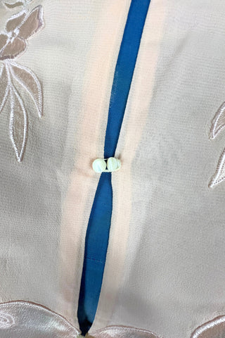 Vintage 1930s silk bed jacket with mandarin collar in mixed satin and crepe silk, with embroidered appliqué flowers and swirls. The three-quarter length sleeves have wide folded cuffs embellished with matching embroidered swirls. Closes in the front with three sets of tiny shiny buttons that resemble ivory.

Label reads Classic Silk Underwear. Made in Canada. Size Small.
