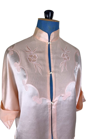 Vintage 1930s silk bed jacket with mandarin collar in mixed satin and crepe silk, with embroidered appliqué flowers and swirls. The three-quarter length sleeves have wide folded cuffs embellished with matching embroidered swirls. Closes in the front with three sets of tiny shiny buttons that resemble ivory.

Label reads Classic Silk Underwear. Made in Canada. Size Small.