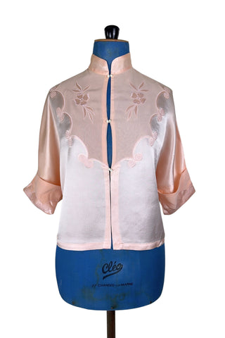Vintage 1930s silk bed jacket with mandarin collar in mixed satin and crepe silk, with embroidered appliqué flowers and swirls. The three-quarter length sleeves have wide folded cuffs embellished with matching embroidered swirls. Closes in the front with three sets of tiny shiny buttons that resemble ivory.

Label reads Classic Silk Underwear. Made in Canada. Size Small.