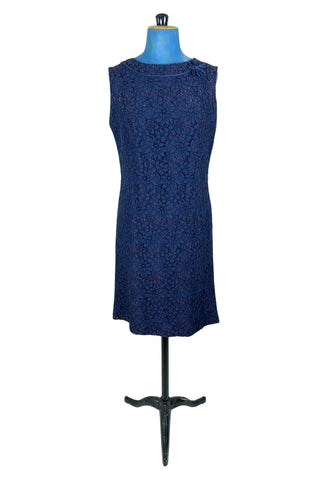 Vintage 1950s cocktail dress in navy blue floral lace, featuring open corded lace over a contrasting mauve lining. The sleeveless sheath dress has a boatneck neckline trimmed with navy cord which is gathered into a flower brooch. The dress closes in the back with a cotton twill zipper.