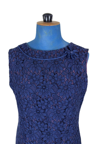 Vintage 1950s cocktail dress in navy blue floral lace, featuring open corded lace over a contrasting mauve lining. The sleeveless sheath dress has a boatneck neckline trimmed with navy cord which is gathered into a flower brooch. The dress closes in the back with a cotton twill zipper.