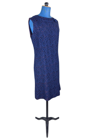 Vintage 1950s cocktail dress in navy blue floral lace, featuring open corded lace over a contrasting mauve lining. The sleeveless sheath dress has a boatneck neckline trimmed with navy cord which is gathered into a flower brooch. The dress closes in the back with a cotton twill zipper.