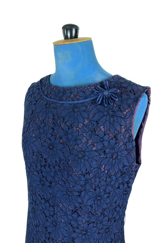 Vintage 1950s cocktail dress in navy blue floral lace, featuring open corded lace over a contrasting mauve lining. The sleeveless sheath dress has a boatneck neckline trimmed with navy cord which is gathered into a flower brooch. The dress closes in the back with a cotton twill zipper.