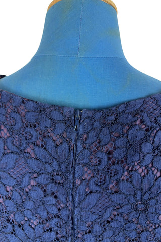 Vintage 1950s cocktail dress in navy blue floral lace, featuring open corded lace over a contrasting mauve lining. The sleeveless sheath dress has a boatneck neckline trimmed with navy cord which is gathered into a flower brooch. The dress closes in the back with a cotton twill zipper.