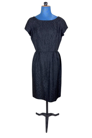 The perfect vintage 1950s little black dress. Perfect for either day or evening wear, with a classic hourglass silhouette and simple round collar and short sleeve sheath design. The skirt is gathered gently into the waist at the hips. The waist is accentuated with matching piping. The dress closes in the back with a metal zipper, and is unlined.

Designed by Roma. Union made by ILGWU AFL-CIO.