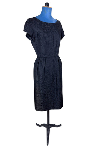 The perfect vintage 1950s little black dress. Perfect for either day or evening wear, with a classic hourglass silhouette and simple round collar and short sleeve sheath design. The skirt is gathered gently into the waist at the hips. The waist is accentuated with matching piping. The dress closes in the back with a metal zipper, and is unlined.

Designed by Roma. Union made by ILGWU AFL-CIO.