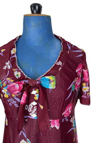 Vintage 1970s dress in burgundy red with a tulip and dianthus floral print. Features short flutter sleeves and a round neckline that can be gathered with the bow tie. The bodice closes with three white diamond shaped plastic buttons. The neckline and sleeves are finished with tiny white scalloped eyelet trim.