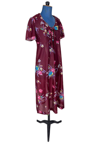 Vintage 1970s dress in burgundy red with a tulip and dianthus floral print. Features short flutter sleeves and a round neckline that can be gathered with the bow tie. The bodice closes with three white diamond shaped plastic buttons. The neckline and sleeves are finished with tiny white scalloped eyelet trim.