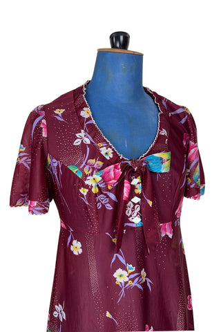 Vintage 1970s dress in burgundy red with a tulip and dianthus floral print. Features short flutter sleeves and a round neckline that can be gathered with the bow tie. The bodice closes with three white diamond shaped plastic buttons. The neckline and sleeves are finished with tiny white scalloped eyelet trim.