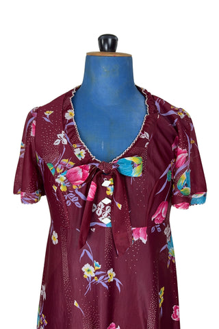 Vintage 1970s dress in burgundy red with a tulip and dianthus floral print. Features short flutter sleeves and a round neckline that can be gathered with the bow tie. The bodice closes with three white diamond shaped plastic buttons. The neckline and sleeves are finished with tiny white scalloped eyelet trim.