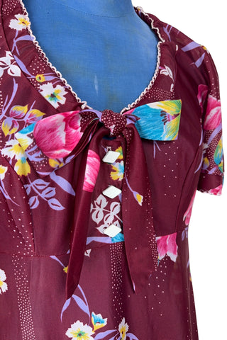 Vintage 1970s dress in burgundy red with a tulip and dianthus floral print. Features short flutter sleeves and a round neckline that can be gathered with the bow tie. The bodice closes with three white diamond shaped plastic buttons. The neckline and sleeves are finished with tiny white scalloped eyelet trim.
