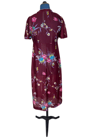 Vintage 1970s dress in burgundy red with a tulip and dianthus floral print. Features short flutter sleeves and a round neckline that can be gathered with the bow tie. The bodice closes with three white diamond shaped plastic buttons. The neckline and sleeves are finished with tiny white scalloped eyelet trim.